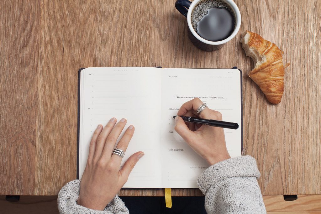 woman writing in journal with a mug of coffee and a croissant on the table