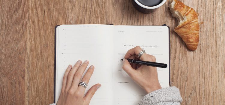 woman writing in journal with a mug of coffee and a croissant on the table