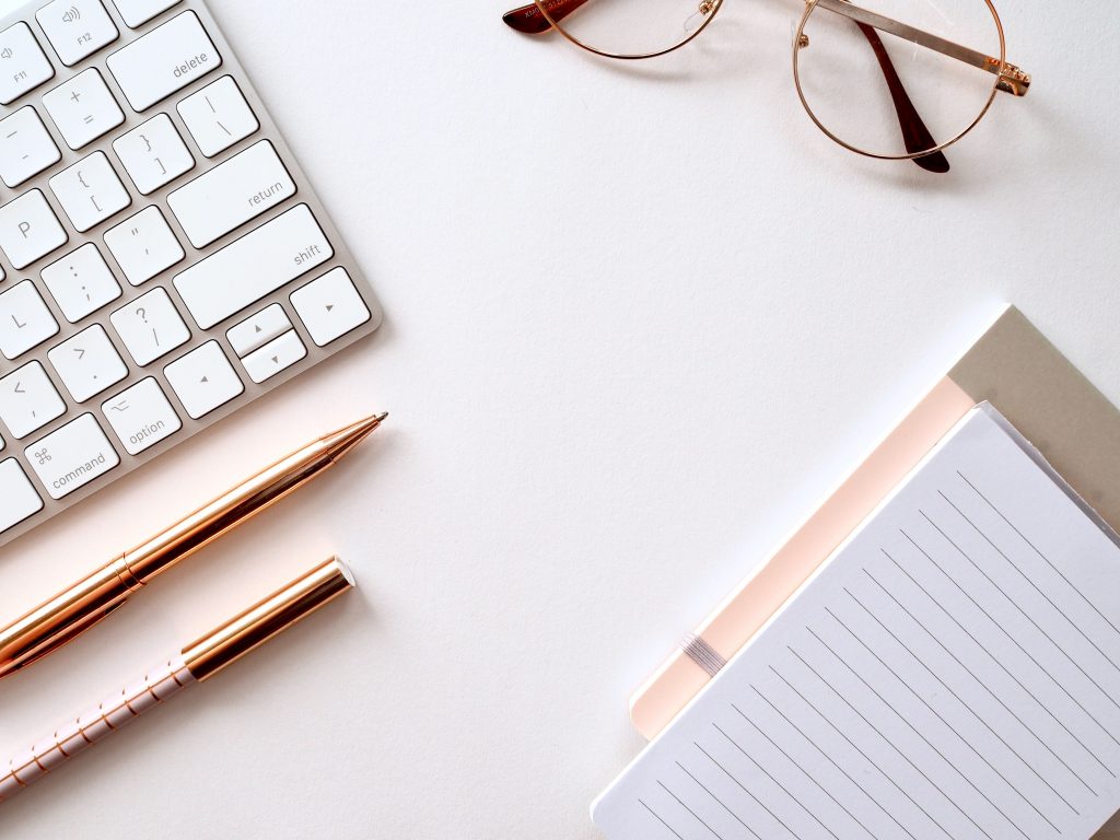 bird's eye view of computer keyboard, notebooks, glasses, and two pens on a white table