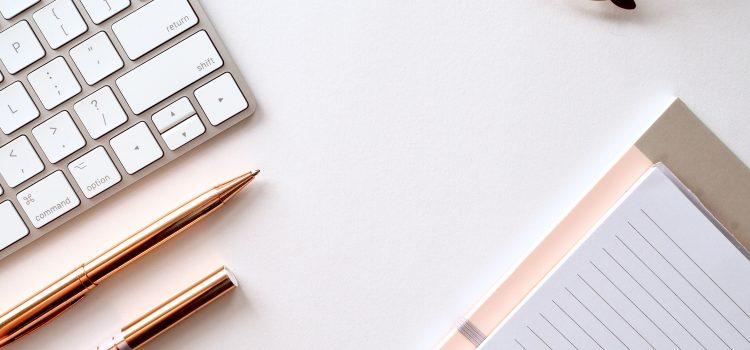 bird's eye view of computer keyboard, notebooks, glasses, and two pens on a white table