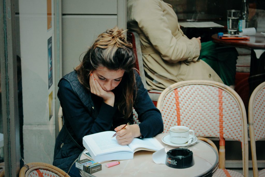 girl reading on a Paris café terrace with a coffee, pack of cigarettes, and ashtray on the table