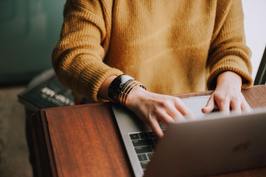 woman wearing mustard yellow sweater typing on a laptop at a wooden table