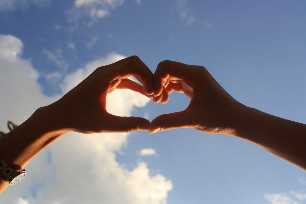 two hands coming together to form a heart shape with a blue sky and some puffy white clouds in the background