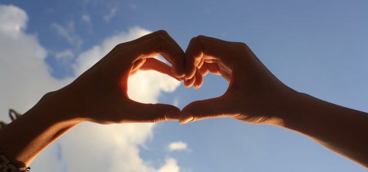 two hands coming together to form a heart shape with a blue sky and some puffy white clouds in the background