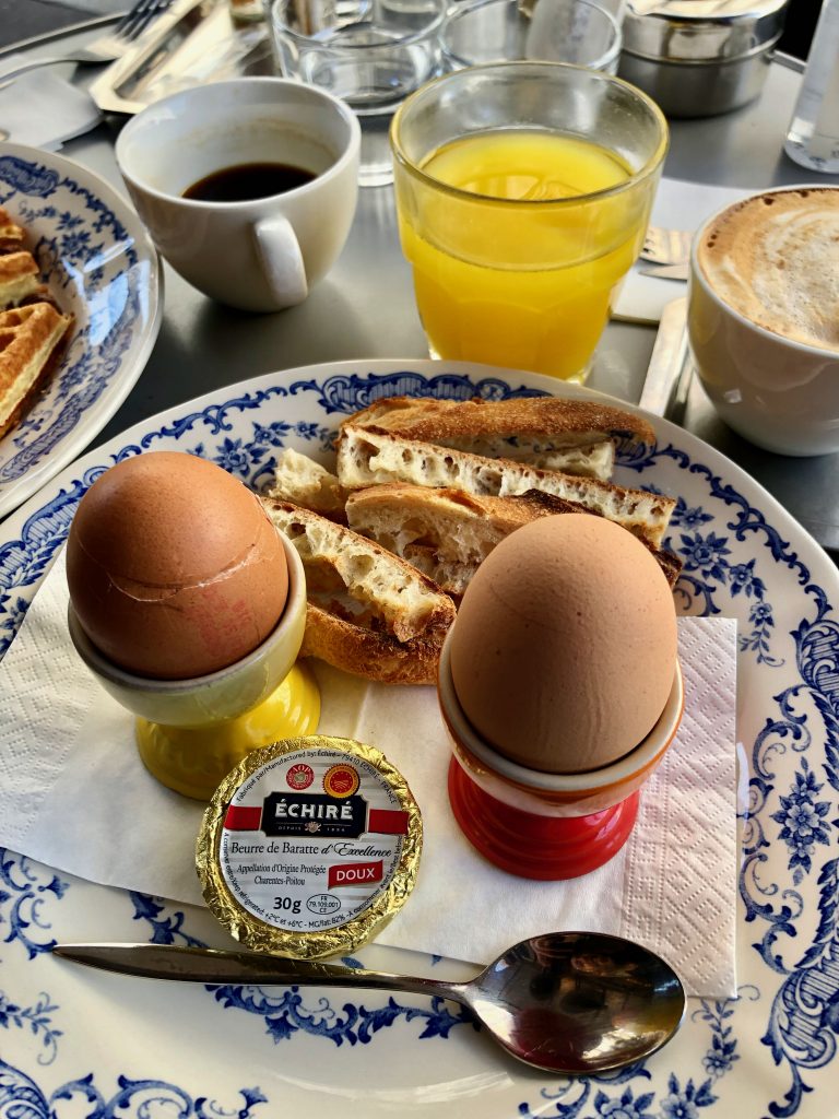 2 œufs à la coque, soft-boiled eggs in egg cups, toasted baguette strips, butter, orange juice, and coffee in a Parisian cafe