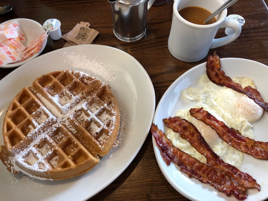 Belgian waffle with powdered sugar, 2 eggs, 4 strips of crispy bacon, and a mug of coffee at an American diner
