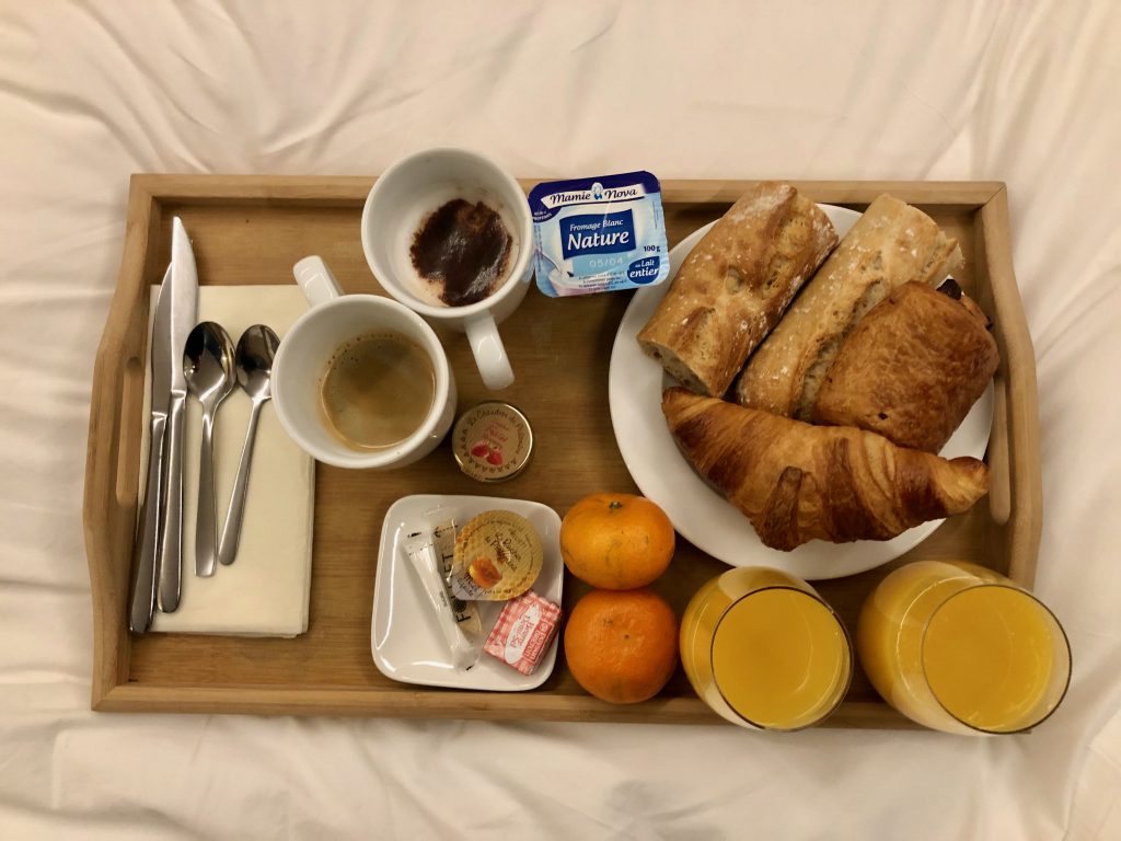 a typical breakfast in France ordered as room service in a French hotel served on a wooden tray: 1 latte and 1 black coffee, 2 glasses of orange juice, 1 croissant, 1 pain au chocolat, slices of baguette (tartines), 1 yogurt, butter, jam, and 2 mandarin oranges