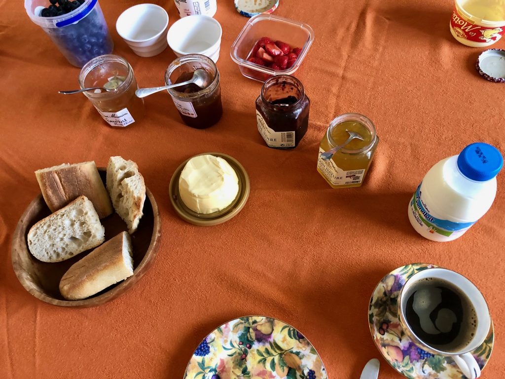 a typical French breakfast laid out on the table: sliced baguette (tartine), butter, jars of jam, fresh fruit (strawberries and blueberries), and coffee