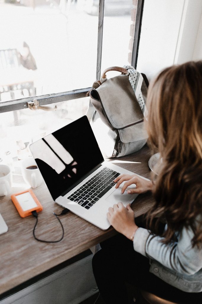 women sitting at a desk, working on a laptop with a coffee nearby and bright light coming in the window