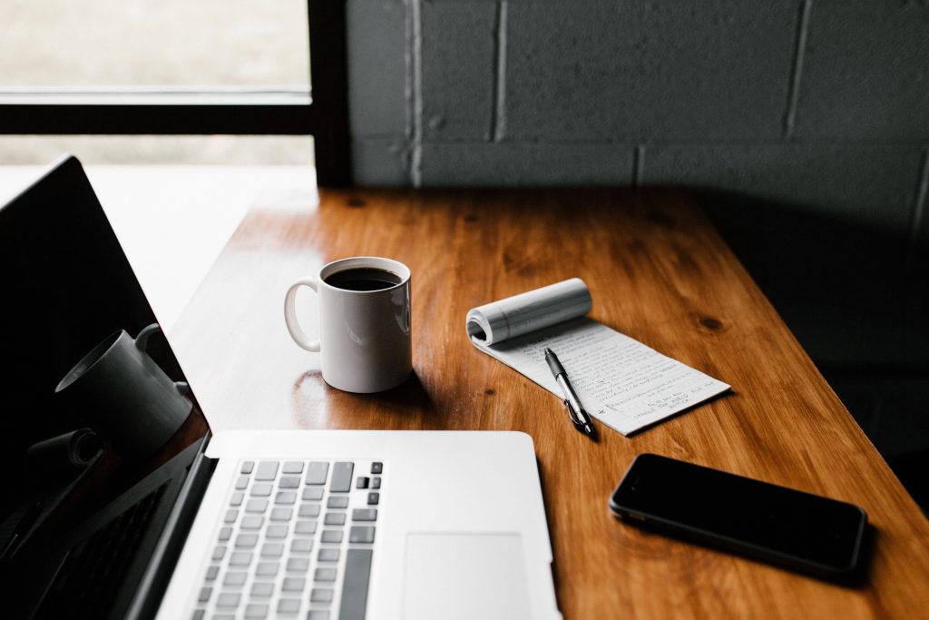 wood table with laptop, mug of black coffee, notepad, pen, and smartphone arranged