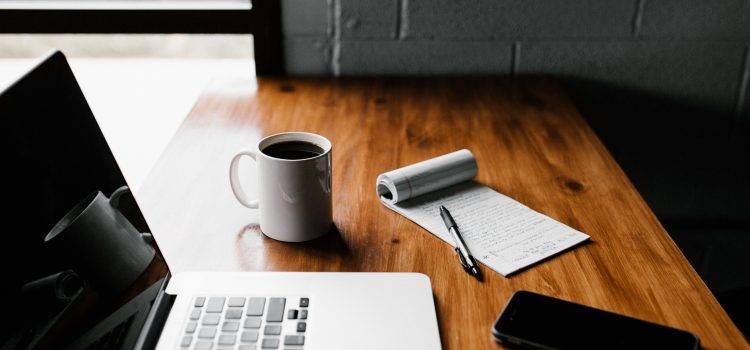 wood table with laptop, mug of black coffee, notepad, pen, and smartphone arranged