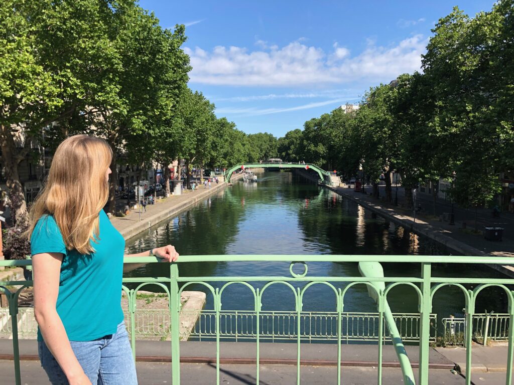 blond woman wearing blue turquoise teeshirt and jeans, looking out over the Canal Saint-Martin on a summer day in Paris