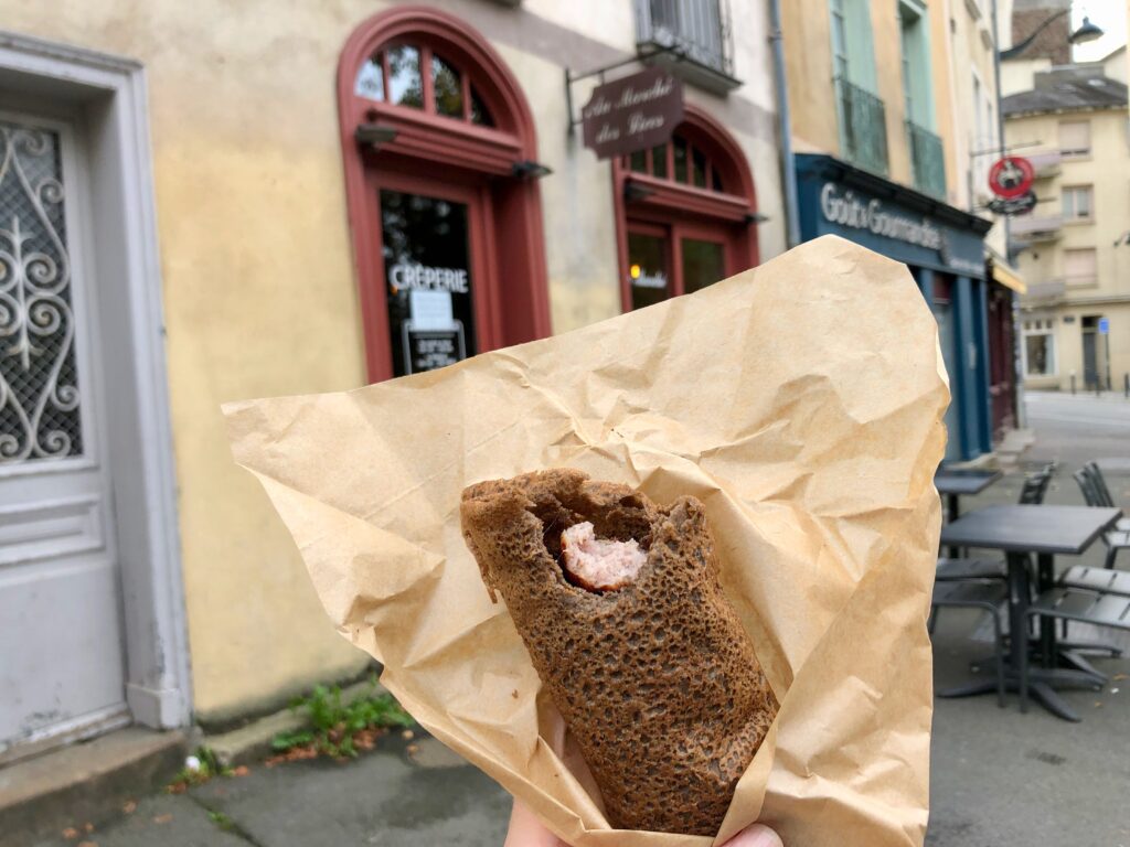 galette saucisse (sausage in a buckwheat flour pancake) wrapped in brown paper and held up with the storefronts in Rennes in the background