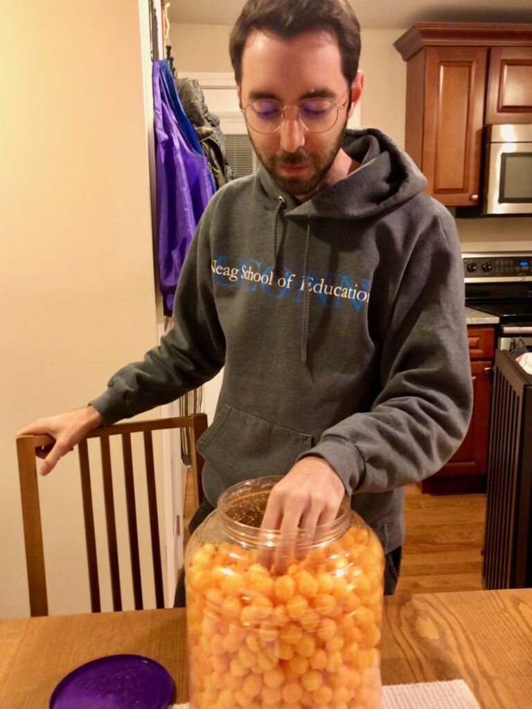 French man wearing a sweatshirt and eating from a large container of cheese balls