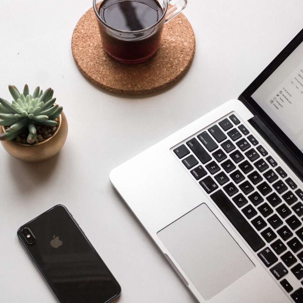 on a white table sits an open laptop, an iPhone, a succulent in a pot, and a glass mug of black coffee