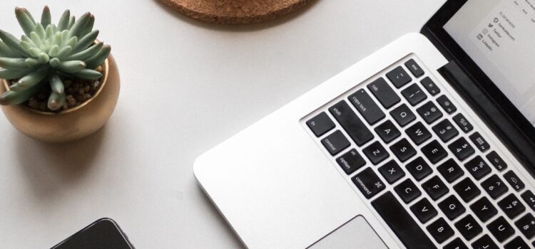 on a white table sits an open laptop, an iPhone, a succulent in a pot, and a glass mug of black coffee