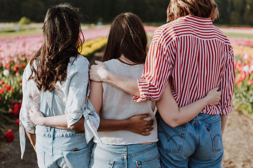 three women standing in a line with their arms around each other and their backs to the camera, staring off into a field of tulips