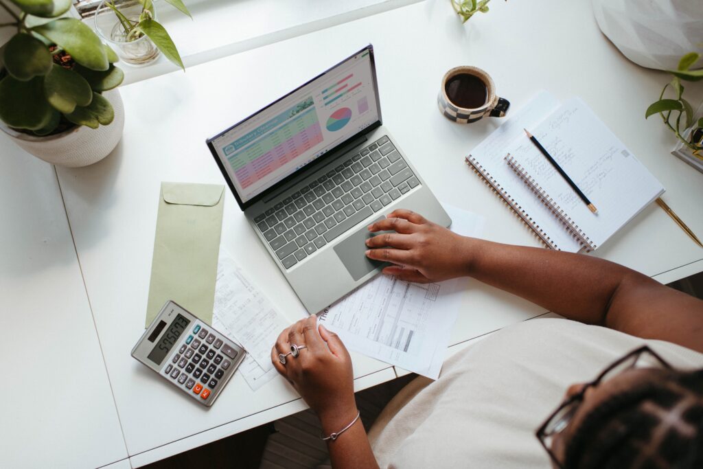 overhead view of a desk with laptop computer, notebooks, calculator, and forms; woman sitting at the desk looking at financial images on computer