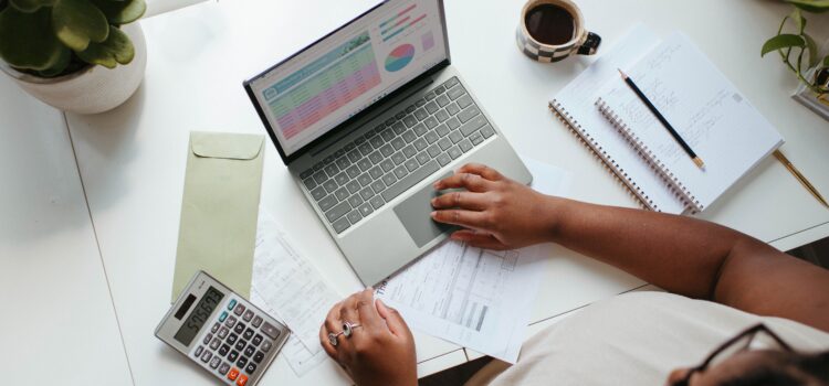 overhead view of a desk with laptop computer, notebooks, calculator, and forms; woman sitting at the desk looking at financial images on computer