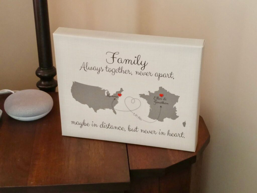 White canvas personalized gift with text: "Family: Always together, never apart, maybe in distance, but never in heart." Below text, on the left, is a gray image of the United States and a red heart on the east coast indicating "Momma." On the right is a gray image of France with a red heart in Paris indicating "Ellen & Jonathan." In between the two hearts is a dotted line indicated that there are 3,547 miles in between these two places.