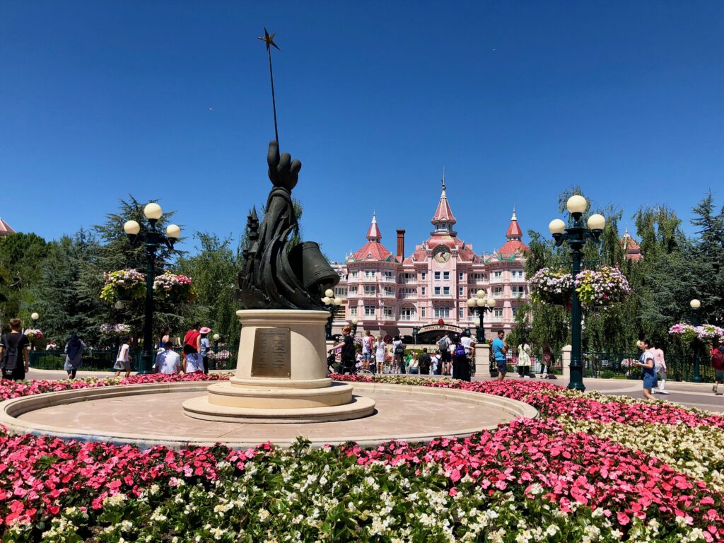 Pink and white flowers in a circular garden with a black statue of a hand holding a magic wand in the middle; Disneyland Paris Hotel in the background