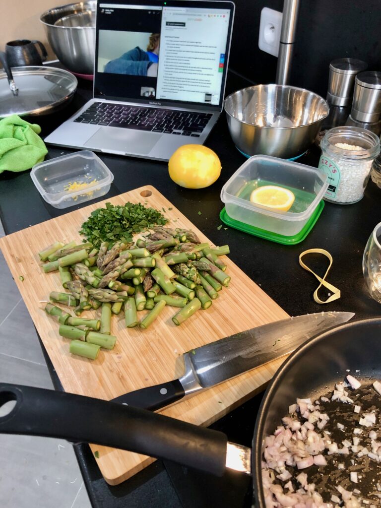 a cooking scene with chopped shallots in a frying pan, chopped asparagus and parsley waiting on a cutting board, a sliced lemon, and a laptop computer open with a videocall and a recipe on the screen for a long-distance Mother's Day celebration