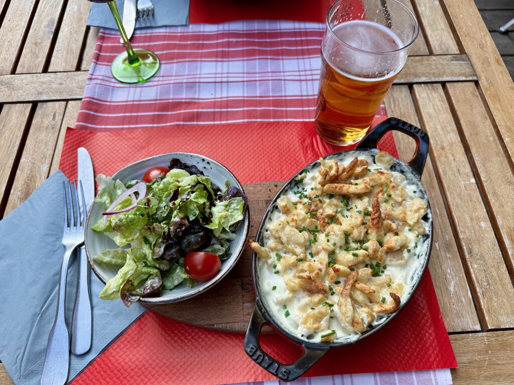 Spätzle au fromage, a traditional Alsatian noodle, served in a cheese sauce in an iron casserole dish. The top of the dish is a little burnt and crispy from the oven. A salad dressed in balsamic vinegar and a large pint of beer are near the main dish.