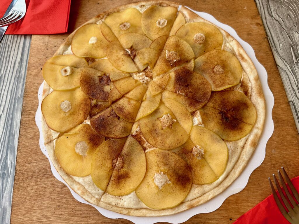 A sweet dessert Flammekueche, also called tarte flambée, on a wooden board with a fork and knife on a red napkin nearby. It’s a very thin and crispy dough rolled out in a circle and topped with thinly sliced apples and cinnamon.