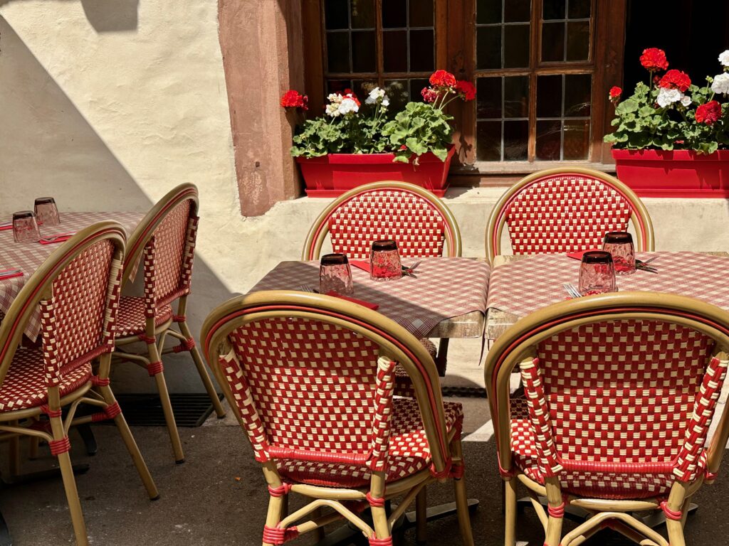 Outside seating area at an Alsatian restaurant in Colmar, France: square tables with red and white checked table clothes, tables are set with glasses and silverware, red and white bistro chairs, and red and white geraniums in a pot on a windowsill behind the tables