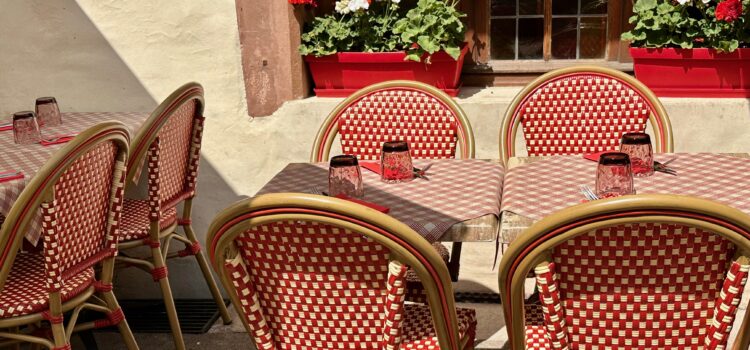Outside seating area at an Alsatian restaurant in Colmar, France: square tables with red and white checked table clothes, tables are set with glasses and silverware, red and white bistro chairs, and red and white geraniums in a pot on a windowsill behind the tables