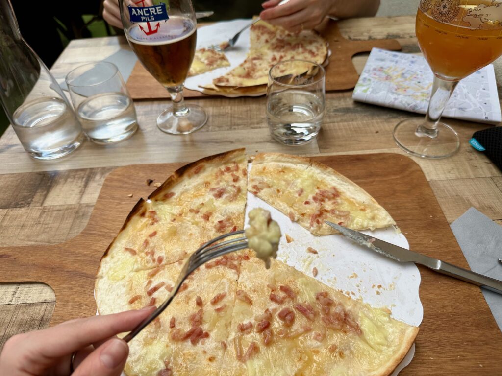 A Flammekueche, also called tarte flambée, on a wooden board being cut and eaten with a fork and knife. It’s a very thin and crispy dough rolled out in a circle and topped with crème fraîche, onions, bacon, and cheese.