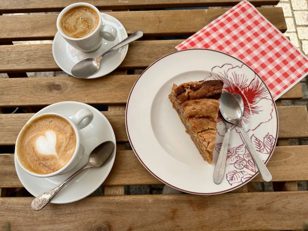 A slice of rhubarb cake on a plate. Two spoons are on the plate and a red and white checked napkin is under the plate. An espresso and a latte, each in their mug on a plate and with a spoon are next to the dessert.