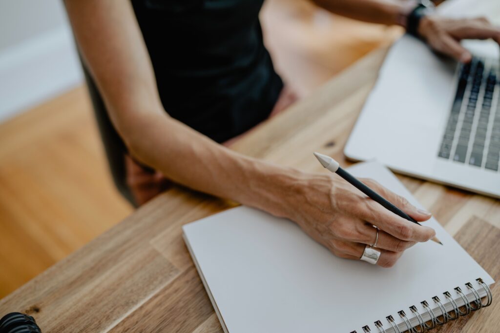 A woman is sitting at a desk. Her right hand is holding a pencil and resting on a blank notebook. Her left hand is hovering over the keyboard of a laptop.