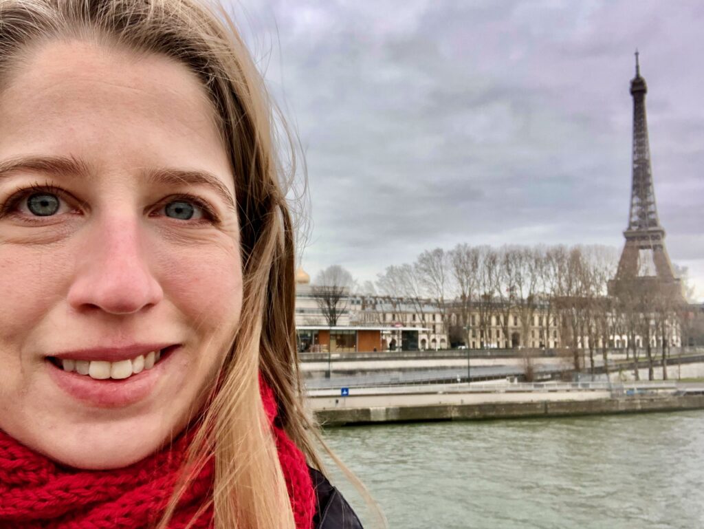 A close-up selfie of a woman wearing a red scarf with the Eiffel Tower in the background. It's winter in Paris and the trees near the tower have no leaves.
