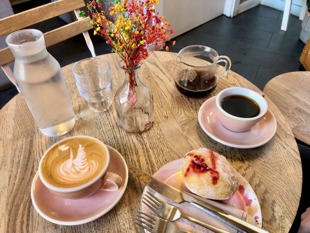 A round wooden table is set with a small vase of dried, colorful fake flowers and a pitcher of water with two small glasses. There is a latte and a brewed coffee in pink mugs on saucers. On a plate is a jam filled donut with two forks and one knife.