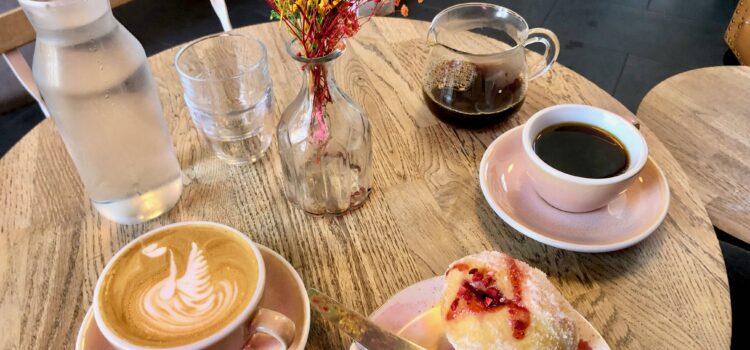 A round wooden table is set with a small vase of dried, colorful fake flowers and a pitcher of water with two small glasses. There is a latte and a brewed coffee in pink mugs on saucers. On a plate is a jam filled donut with two forks and one knife.