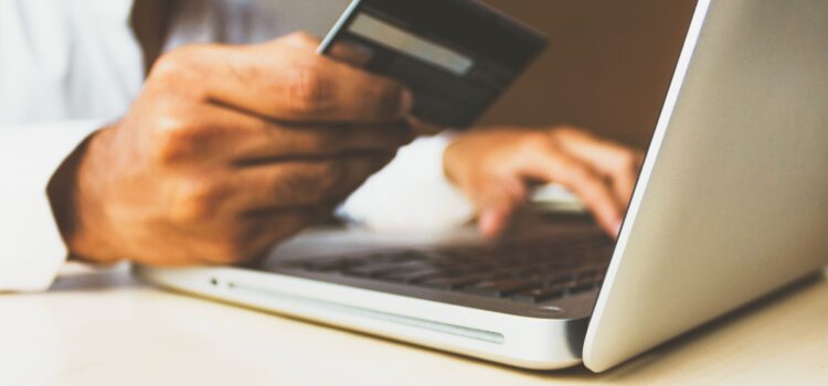 Man making an online payment. He's holding a credit card in his right hand, with his left hand typing on a laptop computer.