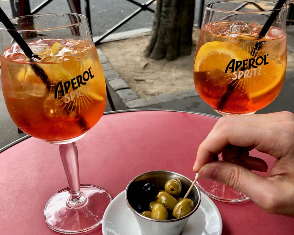 Two full glasses of Aperol Spritz cocktails on a round red table with a small dish of pitted olives in between. A hand is reaching forward with a toothpick to get one of the olives.