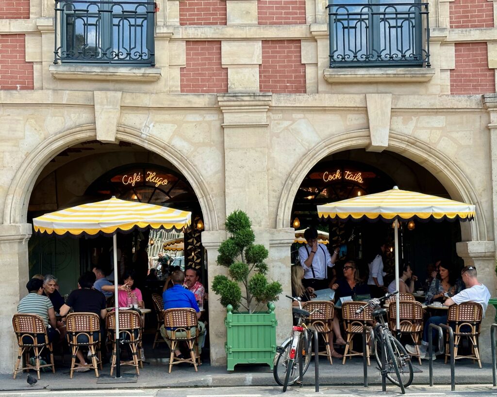 Café Hugo, a well-known café in the Place des Vosges in Paris. Clients are seating at tables nestled under the arcades. Two yellow and white striped umbrellas stand out against the neutral and brick facade.