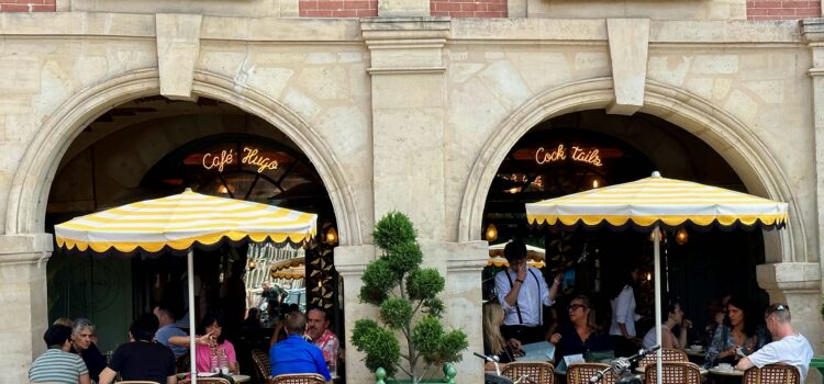 Café Hugo, a well-known café in the Place des Vosges in Paris. Clients are seating at tables nestled under the arcades. Two yellow and white striped umbrellas stand out against the neutral and brick facade.