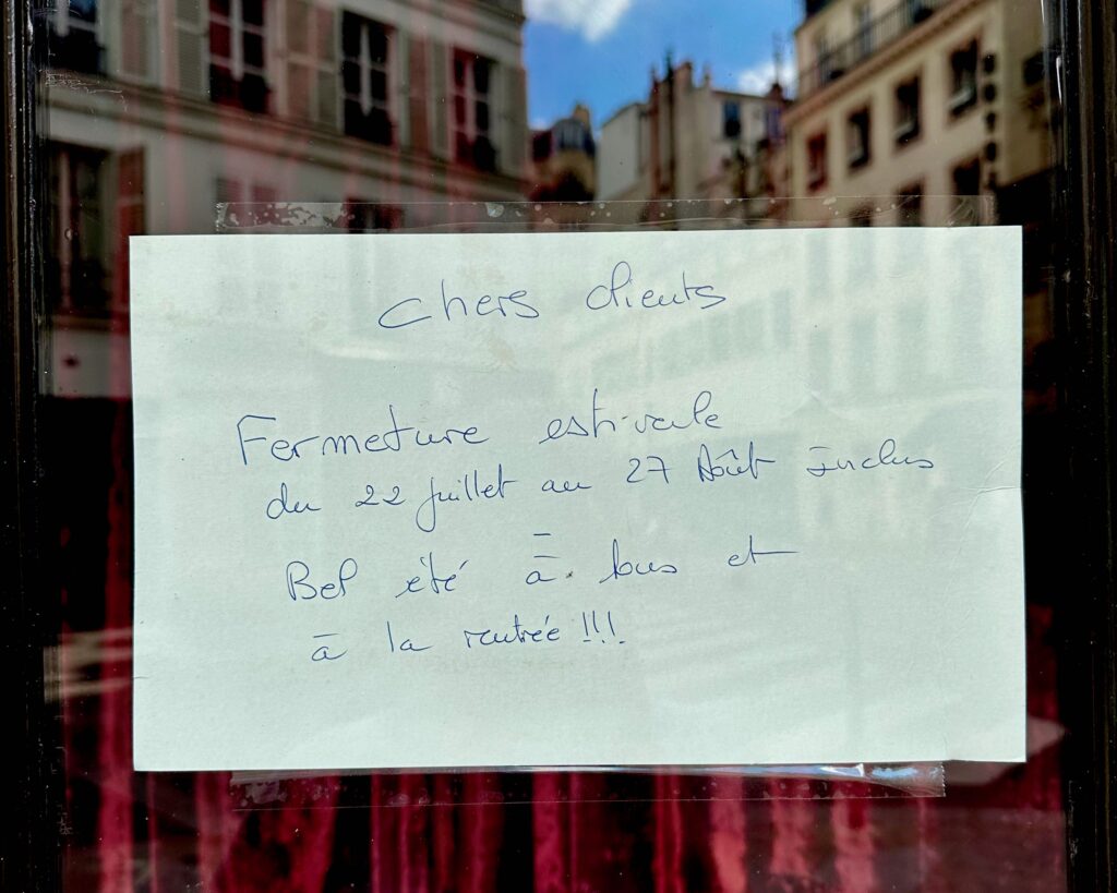 A handwritten sign affixed to the door of a bakery in Paris indicating that they are closed for summer holiday from July 22 to August 27.