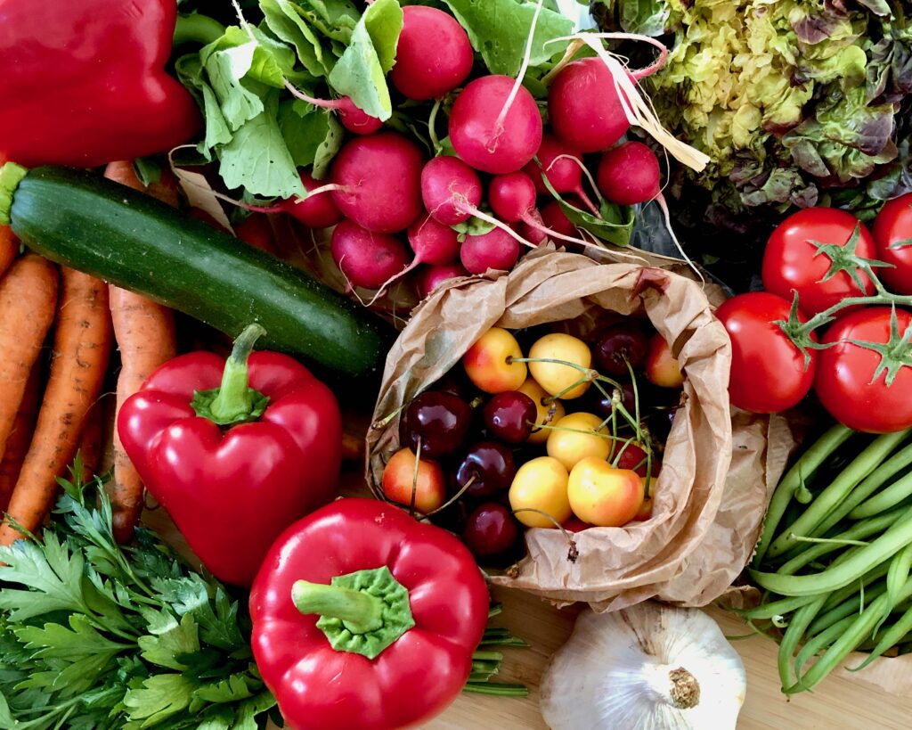 Shopping haul from a trip to the local outdoor food market (marché) in Paris: 3 red bell peppers, a bunch of red radishes, red tomatoes on the vine, a handful of green beans, a bunch of parsley, a few carrots, a head of garlic, a head of lettuce, and a small brown produce bag filled with red and yellow cherries