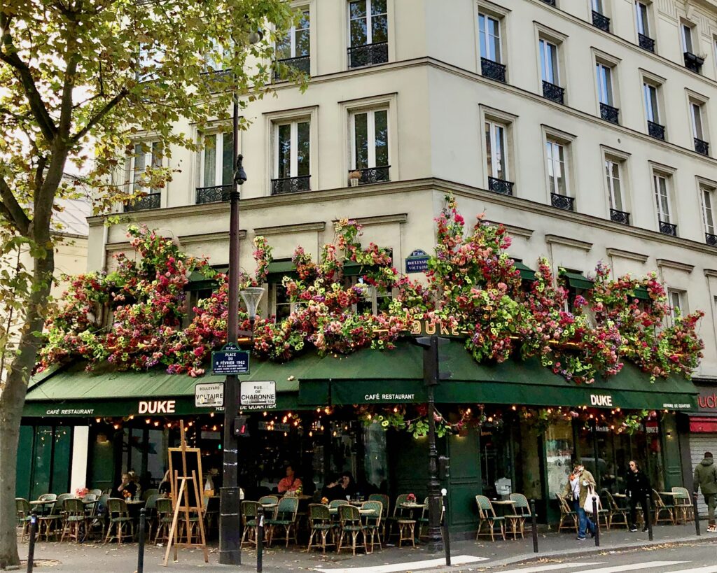 Paris Café with a green awning over the outdoor seating area. Pink and green floral arrangements decorate the awning. Chairs and tables outside with few patrons.