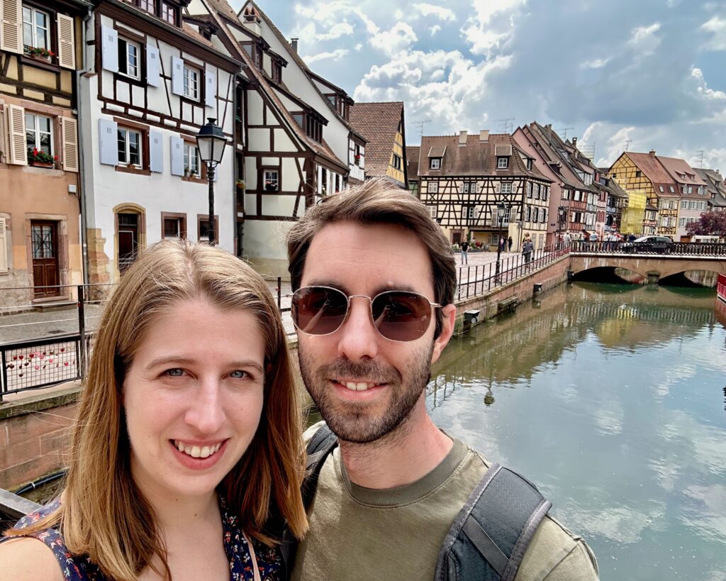 A woman and a man taking a selfie with the colorful half-timbered houses of Colmar, France in the background.