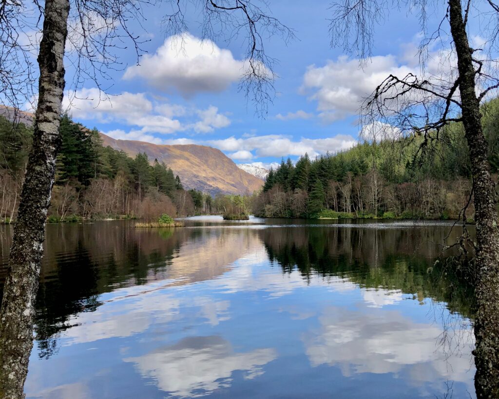 On the Glencoe Lochan Trail in Scotland, a view over a beautiful lake shows a perfect mirrored effect of the tree line and the mountain in the distance. Blue skies and puffy white clouds are as clear in the sky as they are on the lake.