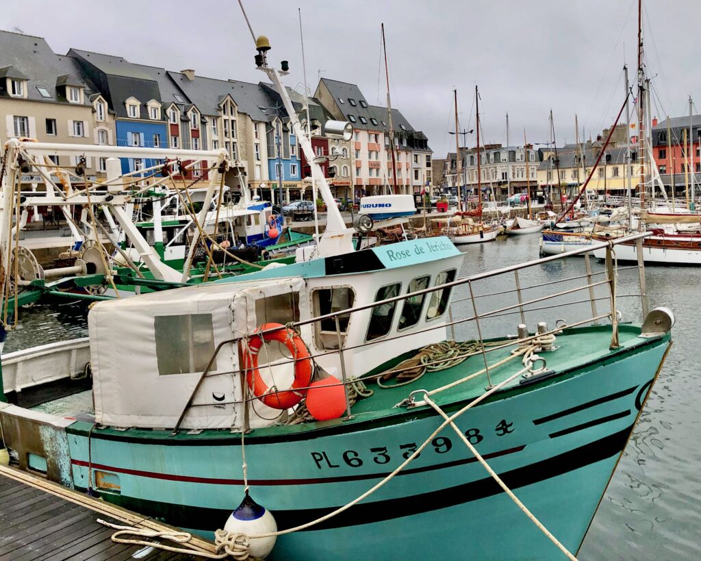 A turquoise and white boat tied to the dock in the fishing port of Paimpol, located in Brittany, France. Colorful houses along the dock and many other boats can be seen in the background.