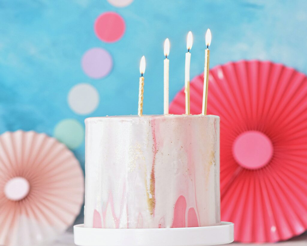 4 candles on a pink birthday cake with white and gold icing dripping down the sides