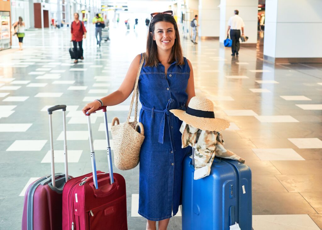 Woman at the airport, prepared for her trip. Smiling and standing with two suitcases to her right and one to her left, sunglasses on her head, and a bag on her shoulder.