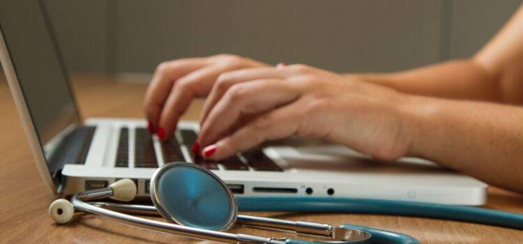 Side view of female doctor's hands placed on laptop computer. Blue stethoscope on the table next to the computer.