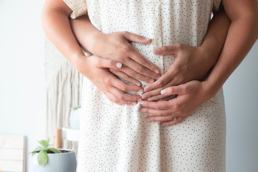 Photo focuses on the mid section of a woman in early pregnancy. Two hands reach around from the back to hold her belly. Her hands are placed on top of theirs.
